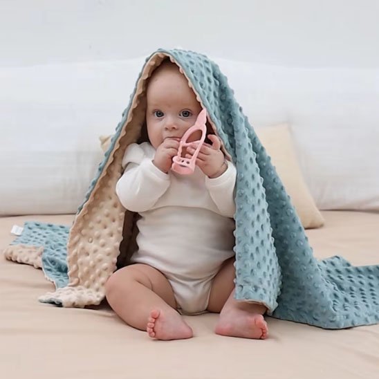 Baby under cozy blue minky blanket holding pink teether, soft sleep accessories for gentle nights