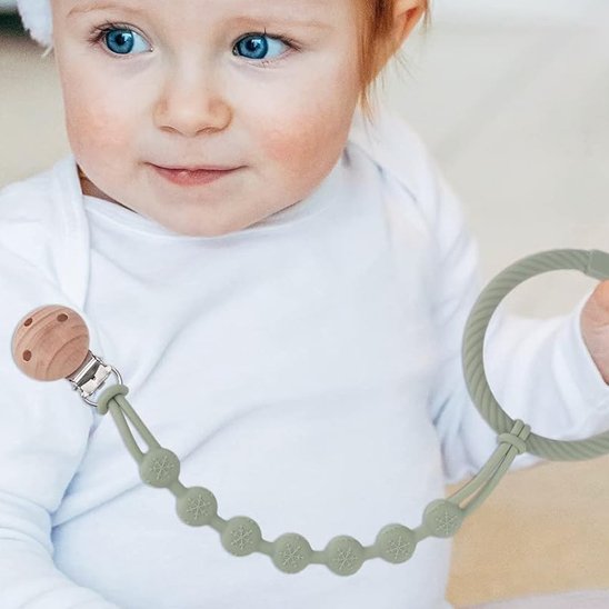 Baby holding olive green teething ring with wooden pacifier clip, soft silicone beads for soothing comfort