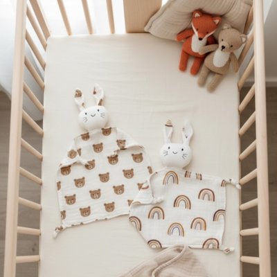 Soft baby comforters with bunny ears and rainbow/bear prints in a wooden crib, perfect for gentle sleep.