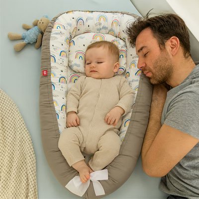 Baby sleeping peacefully in rainbow-patterned bassinet with dad resting nearby, cozy nursery setting