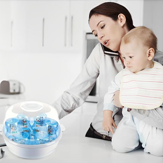 Mother holding baby near baby bottle sterilizer in modern kitchen