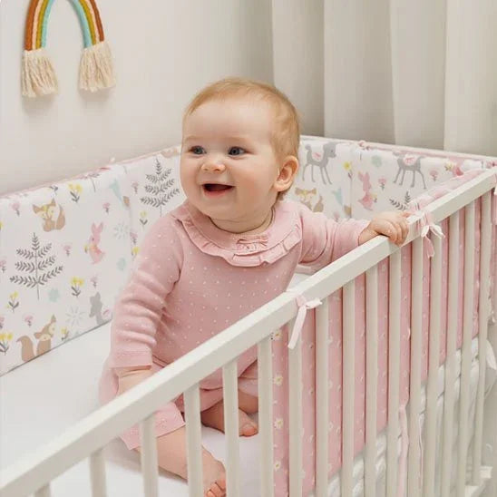 Happy baby in pink pajamas smiling in crib with floral bumper, rainbow wall decor