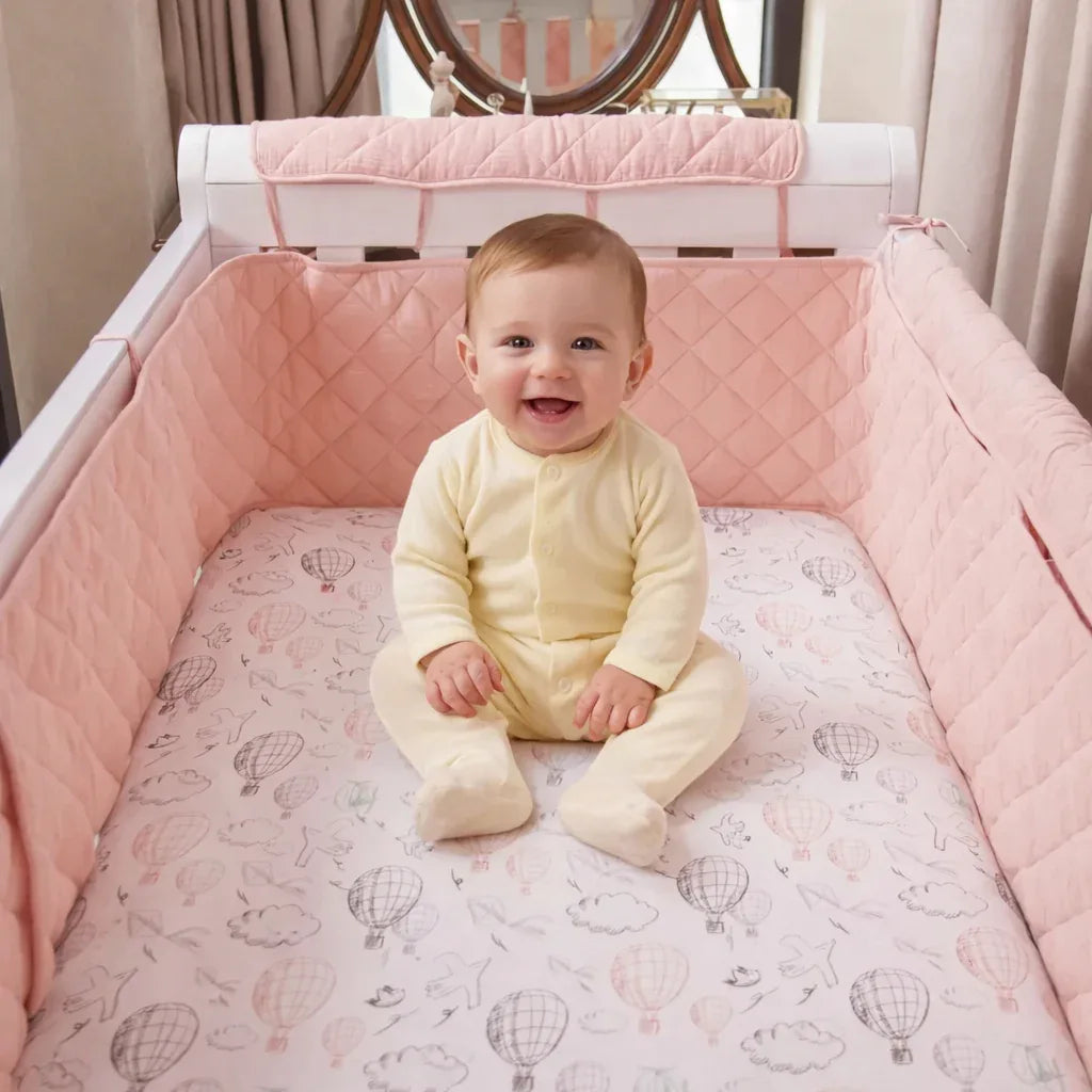 Happy baby in crib with pink quilted bumper and hot air balloon sheet, cozy sleep essentials