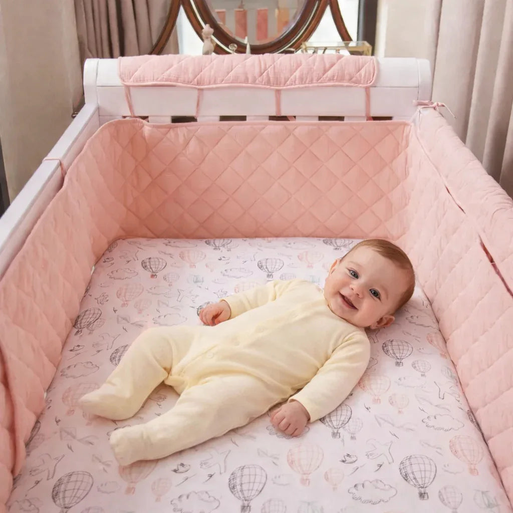Baby smiling in crib with pink quilted bumper and hot air balloon sheet, cozy sleep essentials